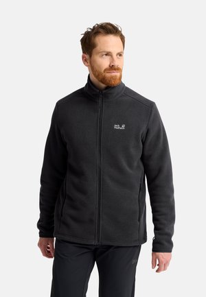 Mann mit rotem Bart, der eine schwarze Fleecejacke mit Reißverschluss und schwarze Hose trägt, schaut leicht seitlich vor einem einfarbigen Hintergrund.
