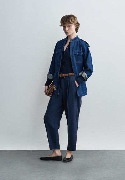 Νεαρό άτομο που φορά μπλε τζιν μπουφάν, σκούρο μπλε πουλόβερ, σκούρο μπλε παντελόνι ψηλόμεσο με καφέ ζώνη, κρατώντας μικρή καφέ τσάντα, φορώντας μαύρα μπαλαρίνες.