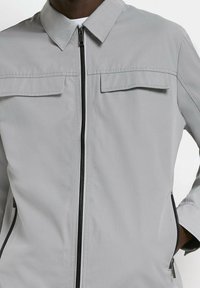 Giacca grigia chiara con colletto a punta, chiusura con zip, due tasche sul petto e zip laterali. Presenta zip nere a contrasto e una superficie in tessuto liscia.
