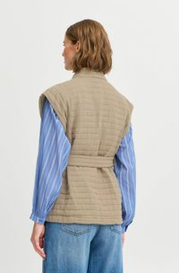 Gilet matelassé beige avec une taille ceinturée, présentant un col haut et des accents d'épaules. Porté sur une chemise à rayures bleues et un jean.