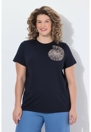 Smilende kvinde med krøllet lysebrunt hår iført en marineblå skjorte med beige blomsterdesign og lyseblå bukser, stående op ad en hvid væg.