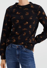 Svart sweatshirt med ett upprepade tigertryck i orange och brunt. Långa ärmar med rund halsringning, tillverkad av ett mjukt material.