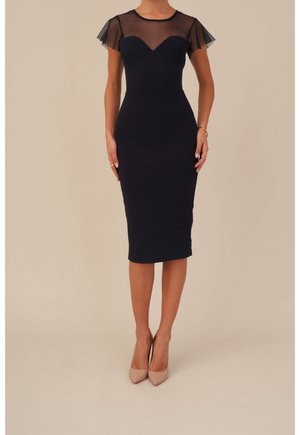 Frau trägt ein eng anliegendes schwarzes Midikleid mit transparenten kurzen Ärmeln und nude High Heels, steht vor einem beigen Hintergrund.