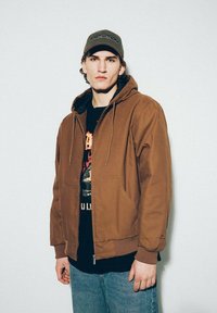 Brązowa kurtka z kapturem, zapinana na zamek, z przednimi kieszeniami, w połączeniu z czarnym t-shirtem z nadrukiem i niebieskimi dżinsami. Akcesorium: zielona czapka.