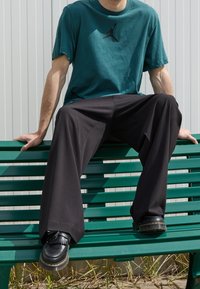 Turkos bomullströja med svart logotyp, kombinerad med svarta vida byxor. Modellen sitter på en grön bänk och har på sig svarta skor med fransar.