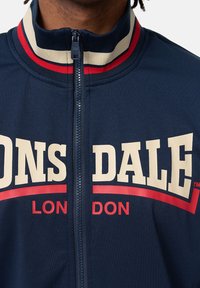 Sötétkék zipzáras dzseki, magas gallérral, piros és krémszínű csíkos részletekkel, valamint feltűnő "Lonsdale London" logóval fehér és piros színben.