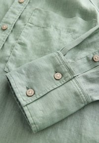 Light green collared shirt in a textured fabric, featuring a chest pocket and beige buttons on the cuffs with visible stitching details.