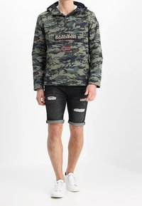 Pullover con motivo camouflato, dotato di cappuccio con cordino e tasca frontale; presenta un'etichetta con logo e una piccola bandiera norvegese. Indossato con shorts neri strappati.