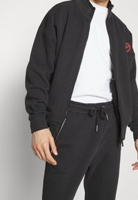 Schwarze Zip-Jacke mit strukturiertem Kragen, ausgestattet mit einem roten Logo und Seitentaschen. Kombiniert mit passenden schwarzen Jogginghosen.