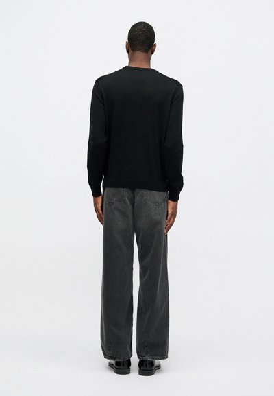 Mann som står med ryggen til, iført en svart langermet skjorte, løse mørkegrå jeans og svarte sko foran en ensfarget hvit bakgrunn.