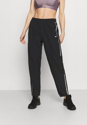 Femme portant un pantalon de jogging noir Nike avec des bandes réfléchissantes, des baskets noires et un soutien-gorge de sport violet, debout devant un fond uni.