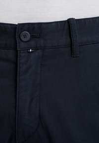 Pantalones azul marino hechos de tela texturizada, con cierre de botón y pasadores para cinturón. Vista en primer plano de la cintura y la zona de la cremallera.