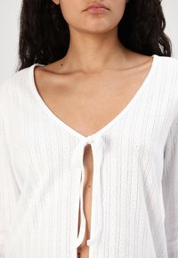 Vit k cardigan med djup V-ringning, med vertikal ribbning och knytning fram, tillverkad av lätt texturerad tyg.