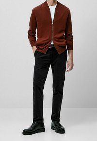 Donkerbruine zip-up cardigan over een witte t-shirt, gecombineerd met zwarte broeken en zwarte veterschoenen. Glad materiaal, getailleerd ontwerp.