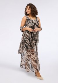 Femme aux cheveux bouclés, portant une robe sans manches à imprimé animal, avec un châle transparent assorti et des talons beiges, se tenant contre un fond clair.