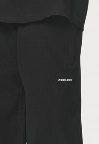 Svarta ribbade byxor i tyg med en sidoficka och en liten vit "PEGADOR"-logotyp broderad på vänster ben.