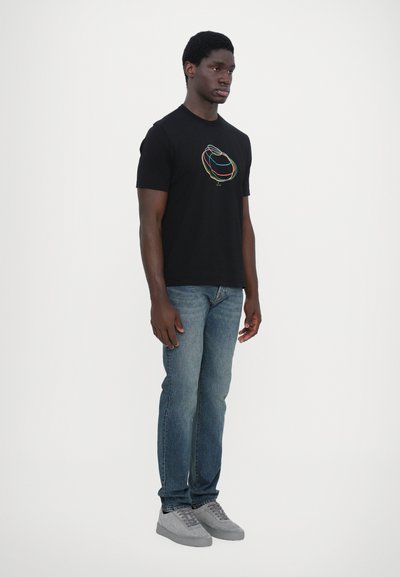 T-shirt en coton noir avec un logo abstrait coloré, associé à un jean en denim bleu clair et des baskets en suède grises. Se tenant devant un fond uni.