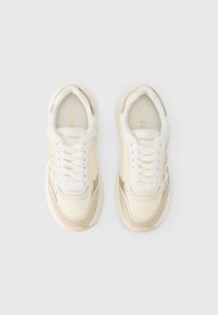 Μπεζ και λευκά αθλητικά παπούτσια με υφασμάτινο επάνω μέρος από μεσοπλέγμα, λείες δερμάτινες λεπτομέρειες, επενδεδυμένο γιακά και διακριτικές χρυσές λεπτομέρειες στα πλάγια.