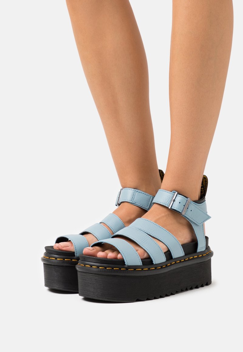 Pieds portant des sandales à plateforme bleu clair à brides, avec des semelles épaisses noires et des coutures jaunes sur un fond clair uni.