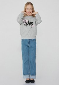 Šedá mikina s mašlí a potiskem "LOVE", zkombinovaná s světle modrými džíny. Oblečení má uvolněný střih a jednoduché černé boty.