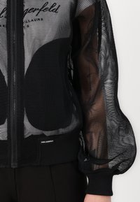 Black mesh jacket featuring a zip closure, puffed sleeves, and a knitted hem. Includes a visible brand label at the lower front.