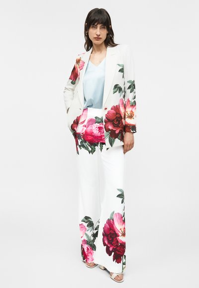 Mujer con traje blanco con grandes estampados florales en rosa y rojo, combinado con blusa azul claro y tacones blancos abiertos, posando contra un fondo blanco.