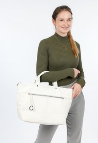 Sac fourre-tout en cuir blanc avec deux poignées courtes, poche zippée à l'avant et détail de pendentif circulaire. Présente une texture lisse et un design minimaliste.