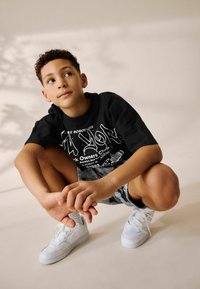 Zwart T-shirt met witte grafische tekst, gestreepte shorts en witte sneakers. Het kind zit in de hurkpositie met de handen op de knieën.