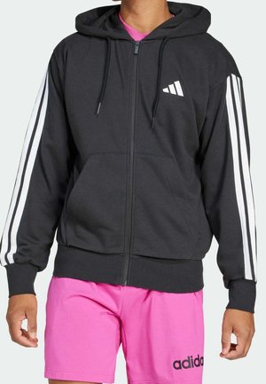 Czarna bluza z zamkiem, białe paski na rękawach i logo Adidas, noszona z jaskrawo różowymi szortami Adidas.