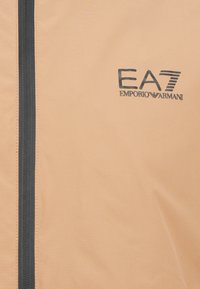 Svijetlosmeđi kaput glatke teksture, s crnim patentnim zatvaračem i crnim logom "EA7 Emporio Armani" otisnutim sprijeda.