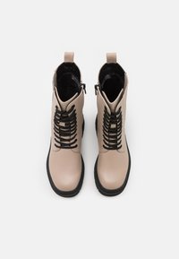 Botine din piele bej, cu sireturi negre și talpă neagră. Prezintă un fermoar lateral și o textură netedă. Design cu vârf rotund.