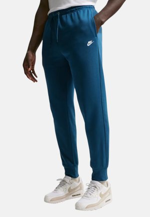 Personne portant un pantalon de jogging bleu Nike et des baskets blanches Nike, debout avec une main dans la poche sur un fond uni.
