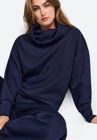 Tamno plavi oversized dukserica s kapuljačom, opuštenog kroja i rebrastim manžetama. Glatka tekstura i minimalni dizajnerski detalji.