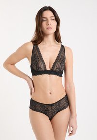 Donna che indossa un bralette in pizzo floreale nero e mutandine coordinate in pizzo, posando con una mano sul fianco e guardando di lato contro uno sfondo bianco.