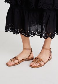 Sandales en cuir brun clair avec une semelle plate, dotées de brides structurées et d'une boucle de cheville réglable, associées à une jupe noire à œillets.