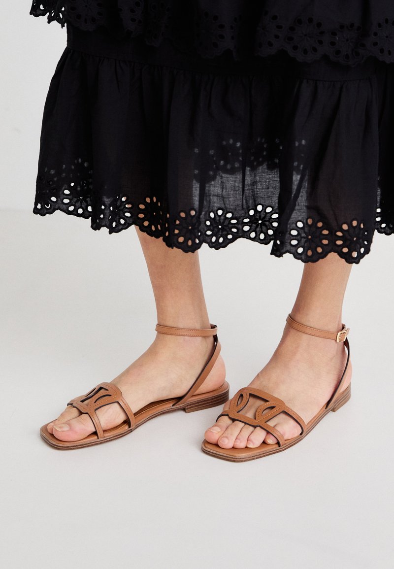 Sandales en cuir brun clair avec une semelle plate, dotées de brides structurées et d'une boucle de cheville réglable, associées à une jupe noire à œillets.