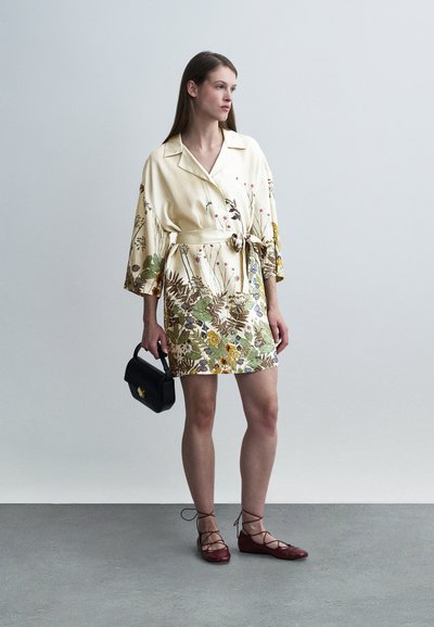 Robe courte fleurie crème avec des motifs verts, jaunes et violets ; ceinture nouée à la taille et manches larges. Sac à main noir, chaussures rouges à lacets.