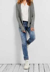 Grå lång cardigan över en vit skjorta med knappar, parat med ljusblå jeans och vita sneakers. Cardiganen har en avslappnad passform och ribbade cuffs.