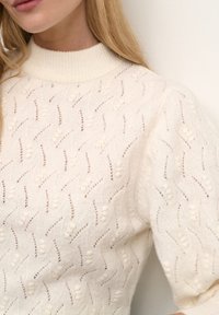 Pull tricoté couleur crème avec un col côtelé haut, présentant un motif en vagues texturé et des accents blancs duveteux.