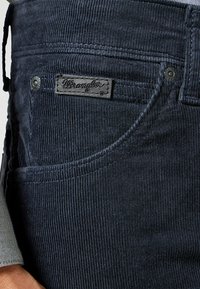 Jeans de pana azul oscuro con una superficie texturizada, que presentan una etiqueta negra de Wrangler en el bolsillo frontal y acentos de metal.