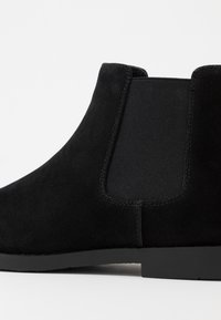 Stivale alla caviglia in suede nero con finitura liscia, pannello laterale elasticizzato e suola piatta in gomma, che mette in risalto un design minimalista e texture.