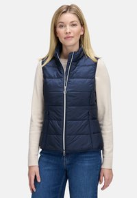 Marineblaues, ärmelloses Steppwesten mit hohem Kragen, gestepptem Design, Vorderreißverschluss und Seitentaschen, getragen über einem hellen Pullover.