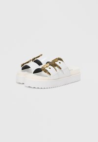 White platform slide sandals with two black straps, gold accent buckles, textured soles, and a smooth finish.