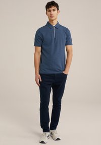 Camisa polo azul marino con una cremallera corta, tela texturada y cuello clásico, combinada con pantalones oscuros y zapatillas ligeras.