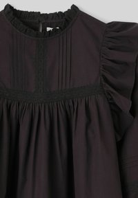 Robe en coton noire avec col et manches à volants. Présente des plis verticaux et une bordure en dentelle le long du corsage et des manches. Silhouette évasée.