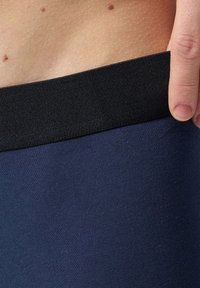 Legging en tissu marine avec une taille élastique noire, texture lisse et coupe ajustée, mettant en valeur la peau au-dessus de la taille.