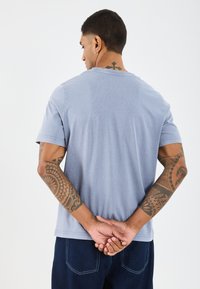 Šviesiai mėlyna trumpomis rankovėmis marškinėliai su apvaliu kaklu, laisvas prigludimas ir tekstūruota medžiaga. Matomi ryškūs tatuiruotės ant rankų. Minimalistinis dizainas.