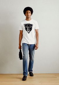 Camiseta blanca con un gráfico de pantera negra, combinada con jeans azules y zapatos negros. El modelo lleva una pequeña bolsa negra.