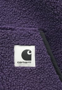 Lillet tekstuuriga fleece-kangas, millel on valge Carhartti logo paan, õmmeldud pinna lähedale musta servaga õmblusele.