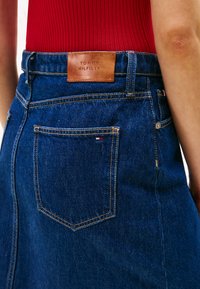 Denimo sijonas su tamsiai mėlynu skalbimu, kuriame yra odinis ženkliukas su išspaustu "TOMMY HILFIGER" ir mažas prekės ženklo etiketas ant nugarinės kišenės.